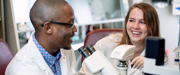 Two members of the lab talking as they work at microscopes