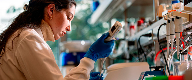 Photograph of the upper torso of a woman in a white lab coat and blue lab gloves sitting in a lab using equipment