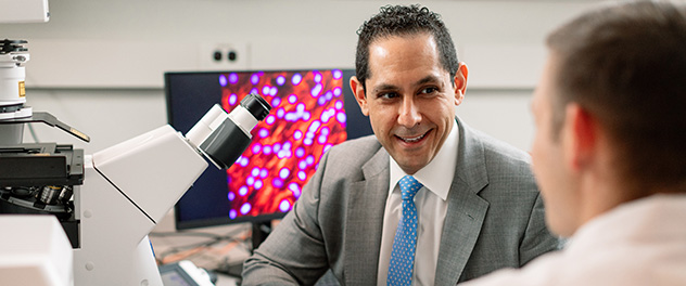 Photograph showing upper torso of Dr. Abdel in a suit and blue tie sitting in front of a microscope talking to another lab worker with his back to the camera