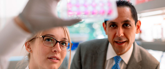 Photograph of the upper torso of Dr. Abdel in a suit and blue tie as he speaks to a woman with glasses working in the lab holding up a piece of lab equipment.