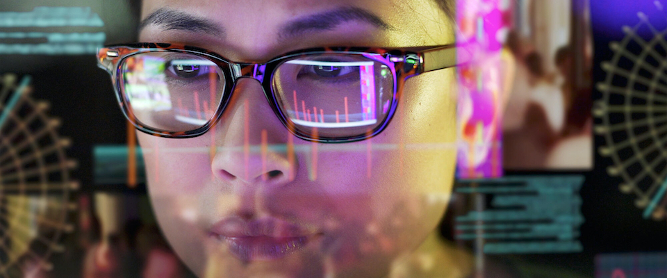 Close-up view of a young woman carefully studying bar graphs and other data on her screen. The screen is transparent, and the camera is looking through the back of the screen.