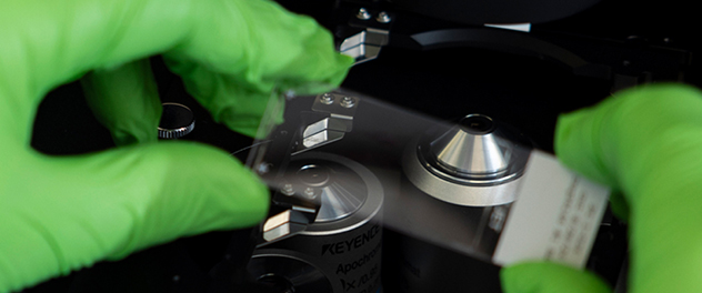 A lab technologist wearing green gloves places a slide into a microscope with the microscope objectives shown in the background.