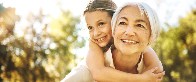 A woman carries her granddaughter on her back outside.