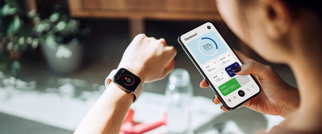 An over-the-shoulder view of a young woman looking at her health data on her smartwatch and smartphone screens.