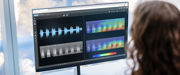 A person views a computer monitor displaying InfinityView, a software interface featuring four panels of audio visualizations. The screens show a waveform, a gray-scale spectrogram and two colorful heat maps with a legend labeled 'Model Attention' showing a gradient from low to high.