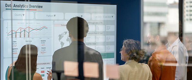 As seen from behind, a group of colleagues view data graphs and charts in a conference room while brainstorming.